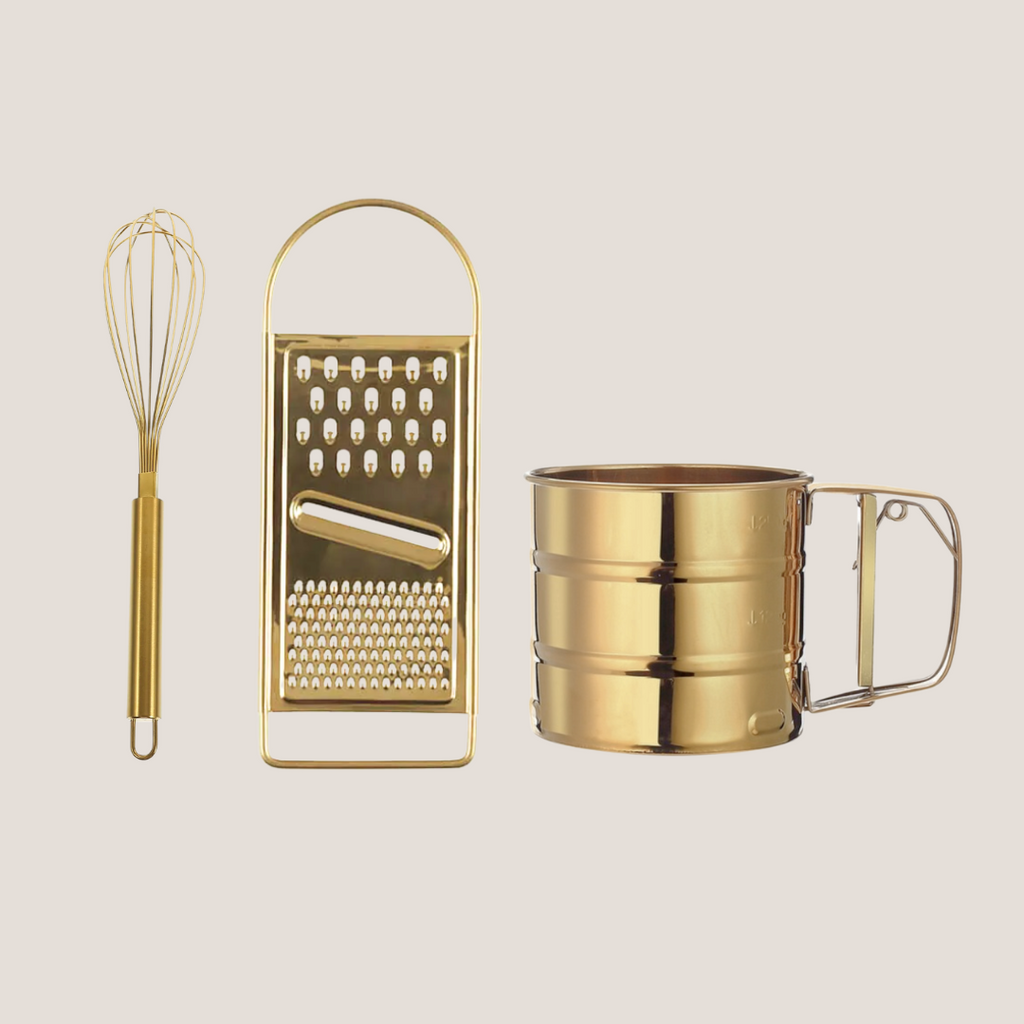 Gold-tone stainless steel egg beater, flour sifter, and grater arranged together on a light background
