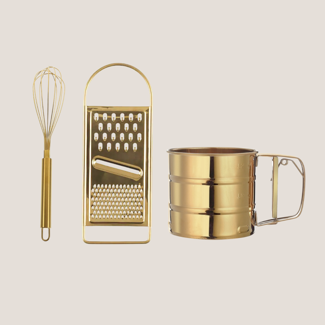 Gold-tone stainless steel egg beater, flour sifter, and grater arranged together on a light background