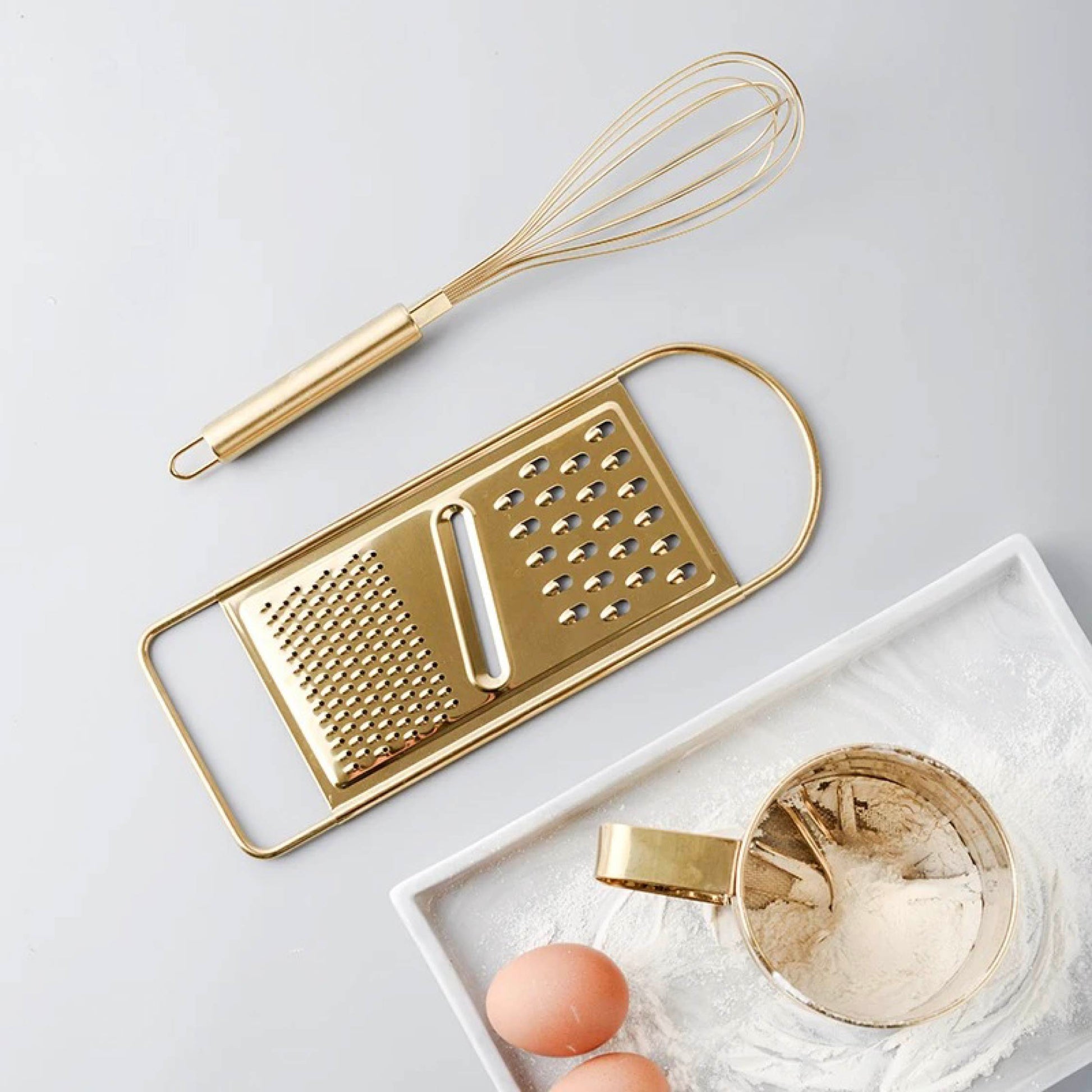 Egg beater, flour sifter, and grater displayed together on a kitchen countertop in a warm lifestyle setting