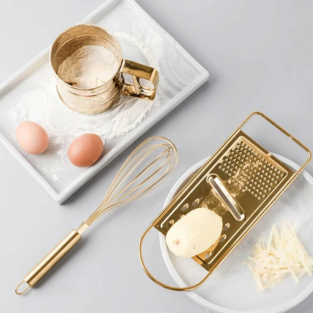 Gold egg beater, sifter, and grater arranged flat on a marble countertop with baking ingredients nearby
